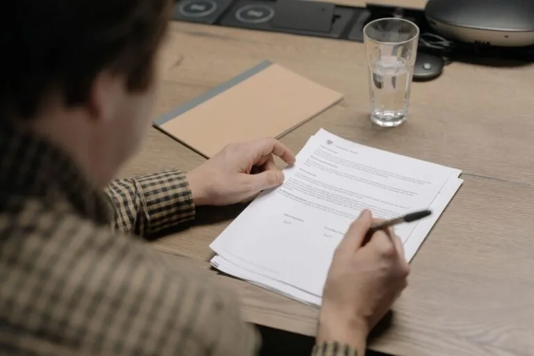 A businessman is reviewing a contract while holding a pen at a desk with a glass of water 