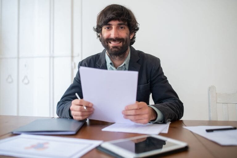 Smiling businessman in a black jacket reviewing documents at his desk, indoors.