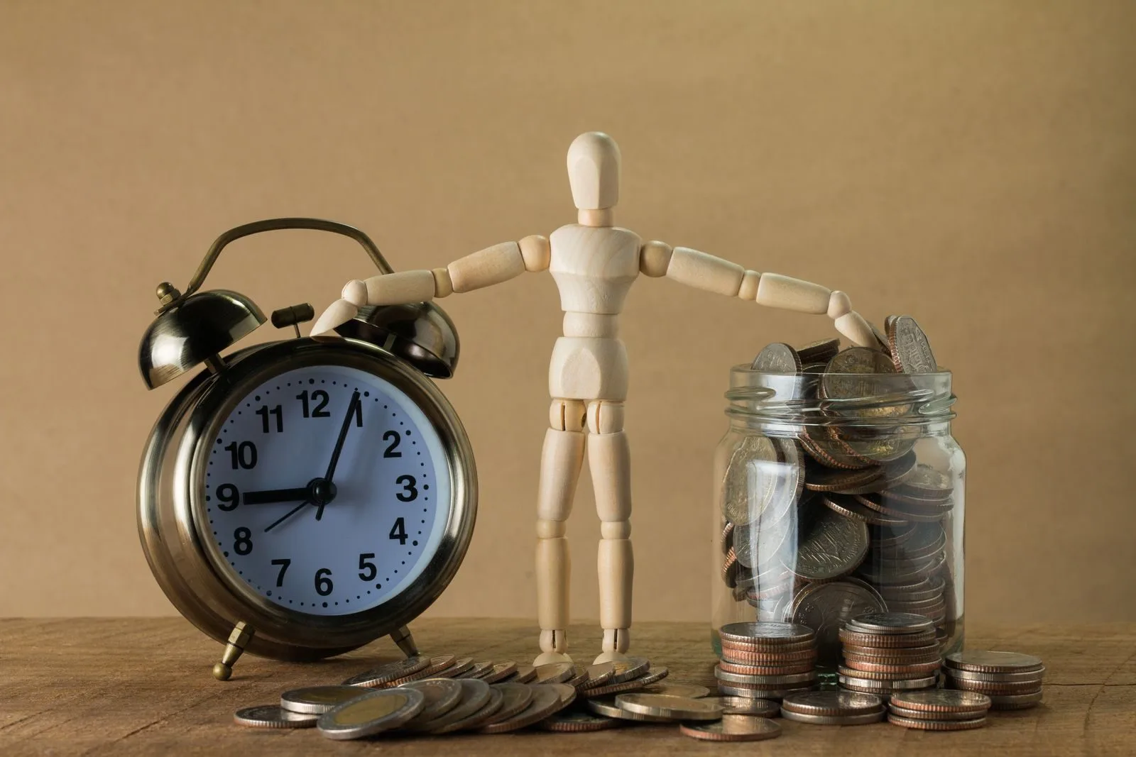 Wooden mannequin holding an alarm clock and a jar full of coins, symbolizing time and money.