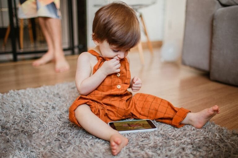 Cute toddler in orange outfit plays with smartphone on carpet indoors.