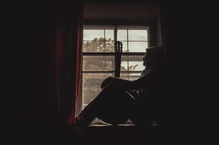 A pensive musician with guitar silhouetted by a window indoors, exuding a moody atmosphere.