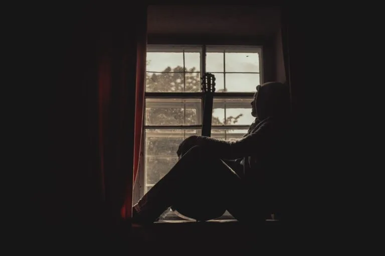 A pensive musician with guitar silhouetted by a window indoors, exuding a moody atmosphere.