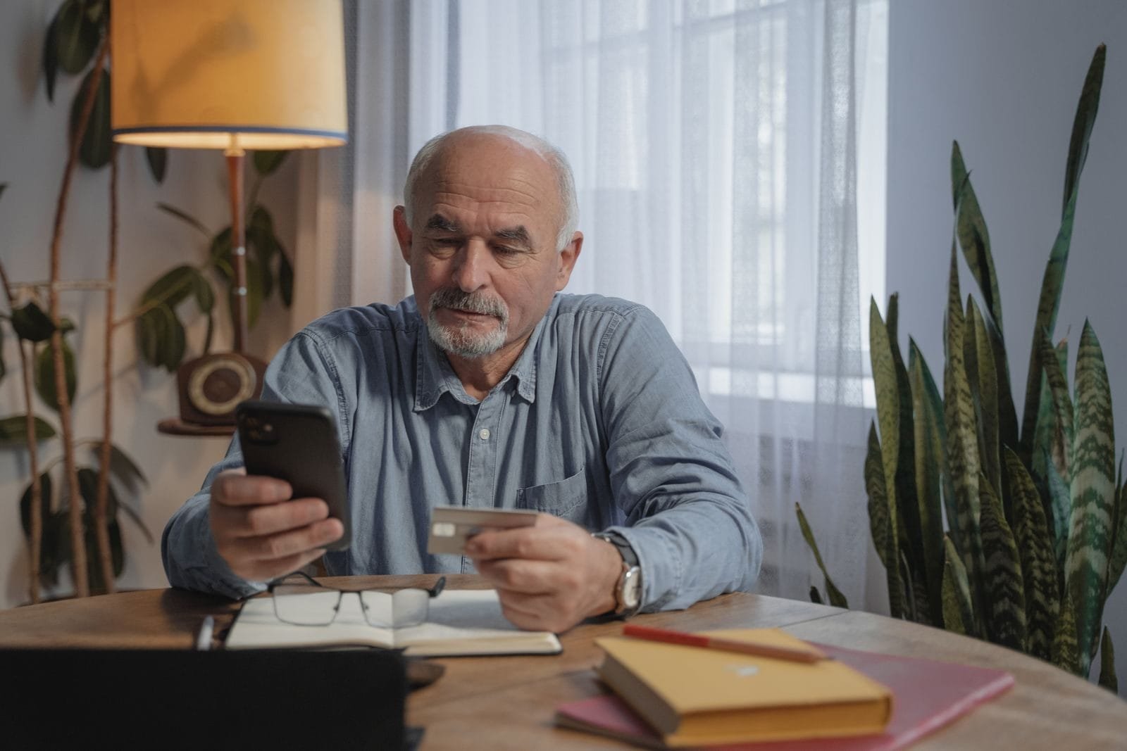 Descontos Associativos: 6,5 Milhões Já Contestaram 1 Elderly man using smartphone for online shopping at home, holding credit card near desk plants.