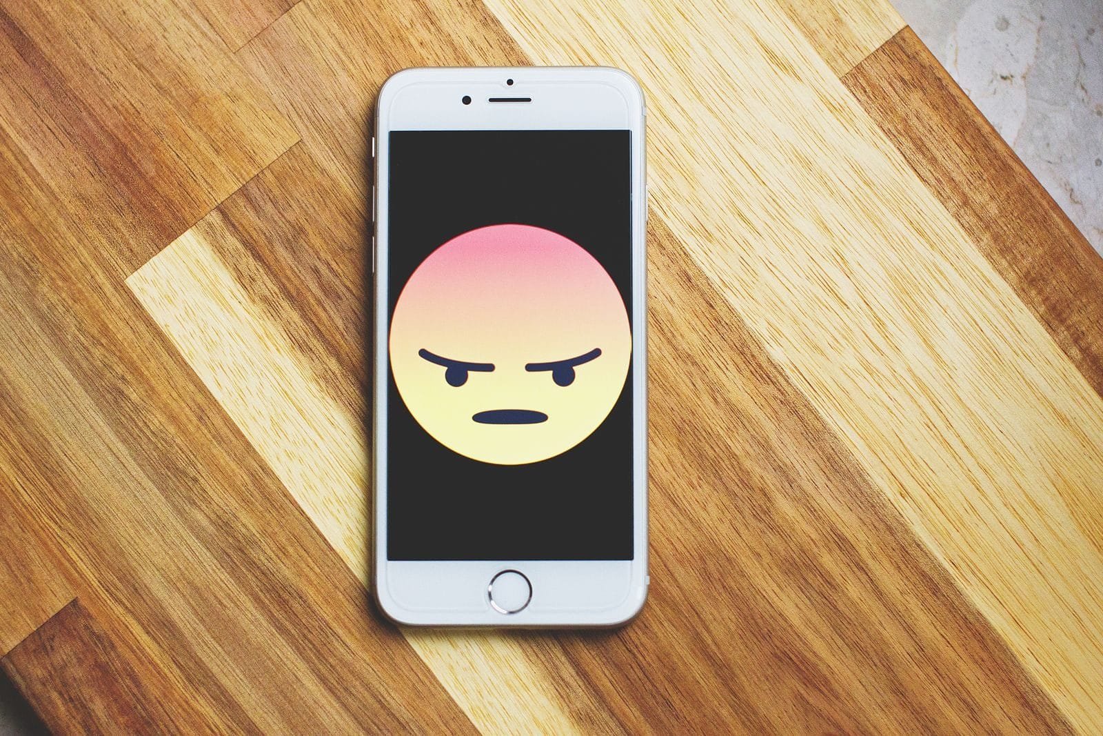 Discurso de ódio e ofensas online: como responsabilizar o autor e a plataforma 1 A smartphone showing an angry emoticon rests on a wooden table, emphasizing tech and emotion.