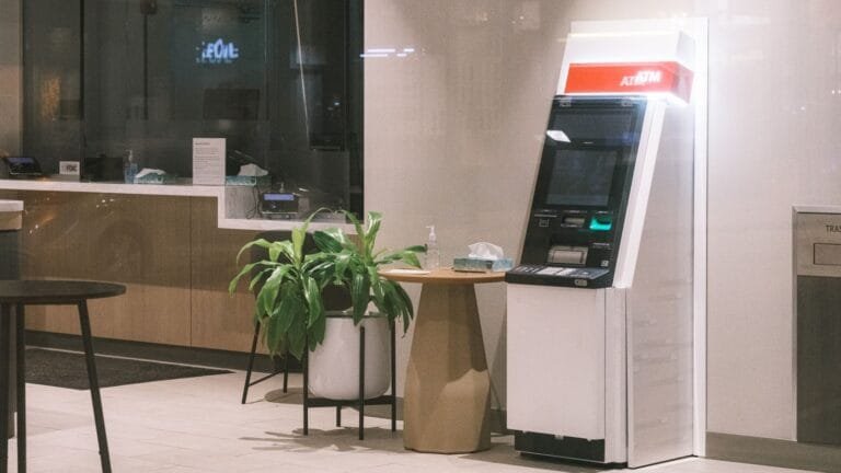 Estabilidade acidentária de doze meses do bancário adoecido 5 An ATM machine stands in a modern bank lobby, next to a plant and table.