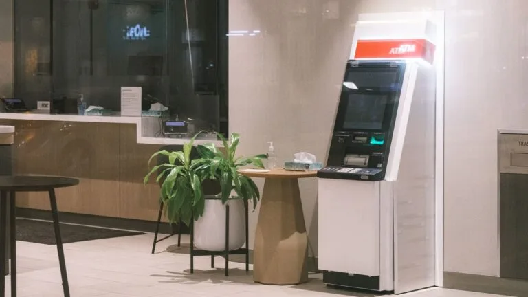 Estabilidade acidentária de doze meses do bancário adoecido 2 An ATM machine stands in a modern bank lobby, next to a plant and table.
