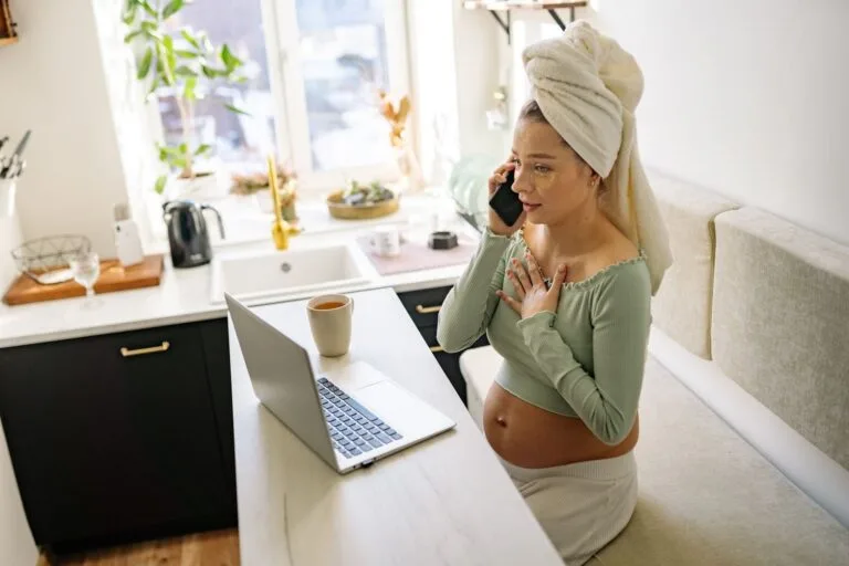 Pregnant woman in casual setting, talking on phone with laptop open and a drink nearby.