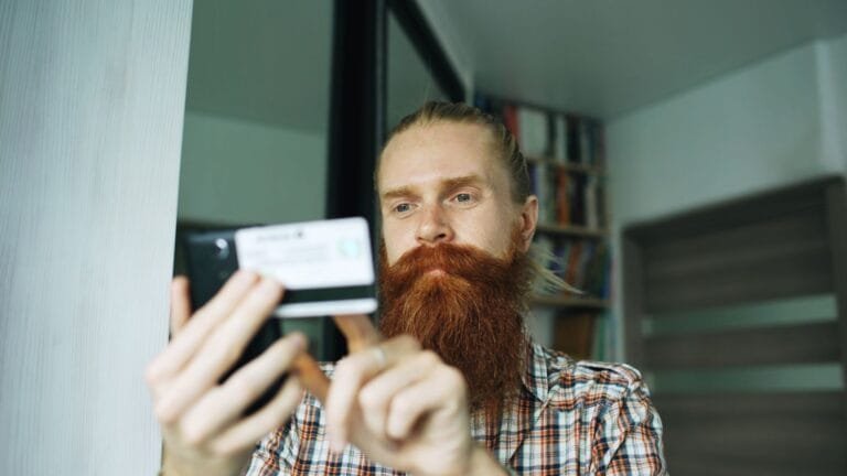 A bearded man uses his smartphone indoors, focusing on a task.