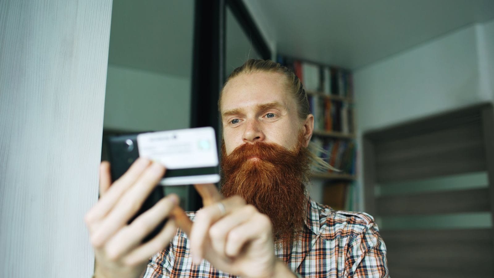 Estelionato no dia a dia: golpes comuns e como buscar reparação 1 A bearded man uses his smartphone indoors, focusing on a task.