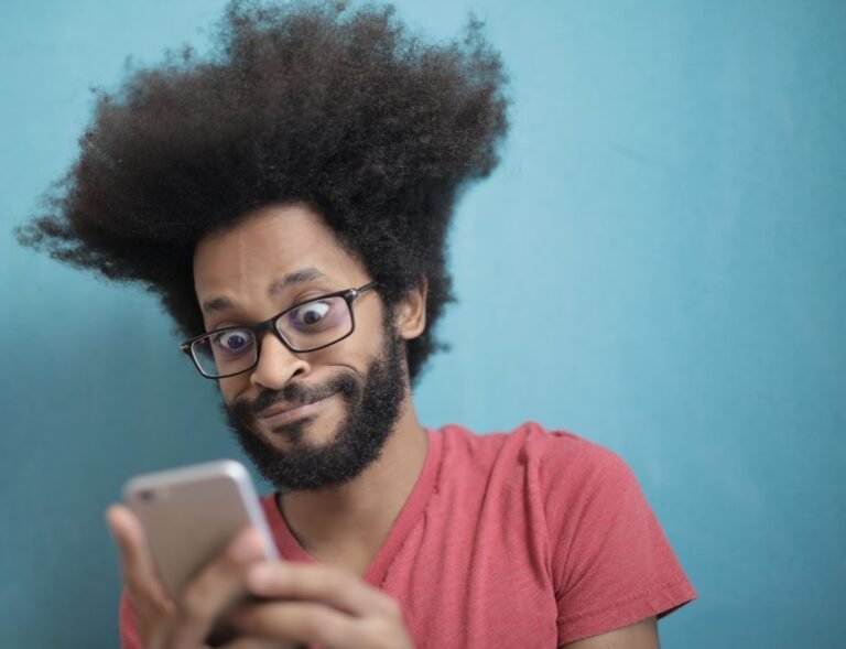 Excited man with afro surprised while texting on smartphone indoors against blue background.