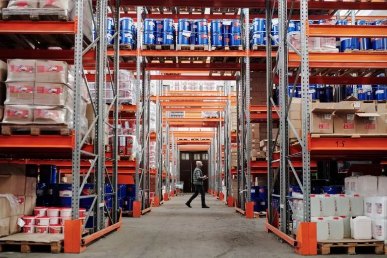 Exclusão do ICMS-ST da Base do PIS/COFINS 7 A man walking through a large industrial warehouse with stacked shelves filled with goods and products.