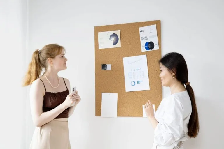 Two women discussing charts on a bulletin board in an office setting.
