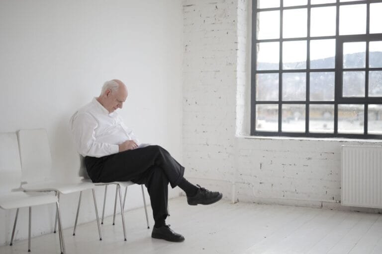 Elderly man in white shirt sitting pensively in a bright, minimalist room with large window.