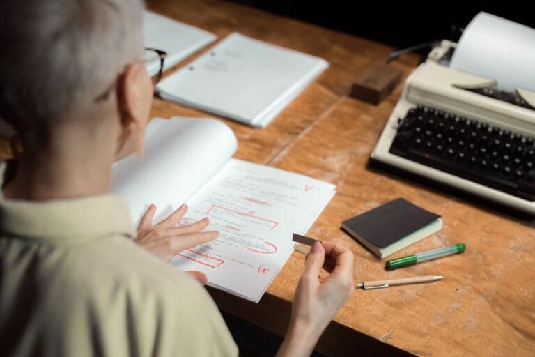 Fila do INSS cai ao menor nível em 21 meses 7 An adult woman reviewing a script with red pen marks at a wooden desk with a typewriter.