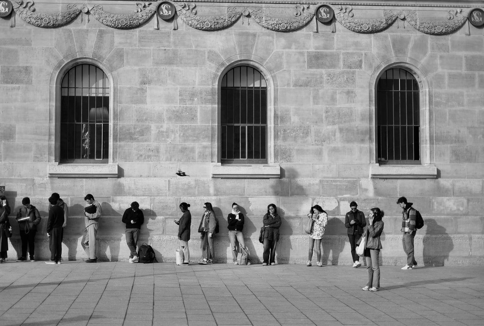 Black and white photo showing a group of people waiting outside a historic stone building with arched windows.