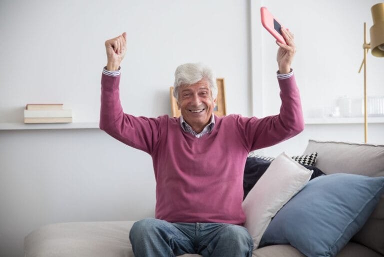Golpe do App Falso do INSS: Como se Proteger em 2026 4 A happy senior man sitting on a sofa holding a smartphone indoors, celebrating with raised arms.