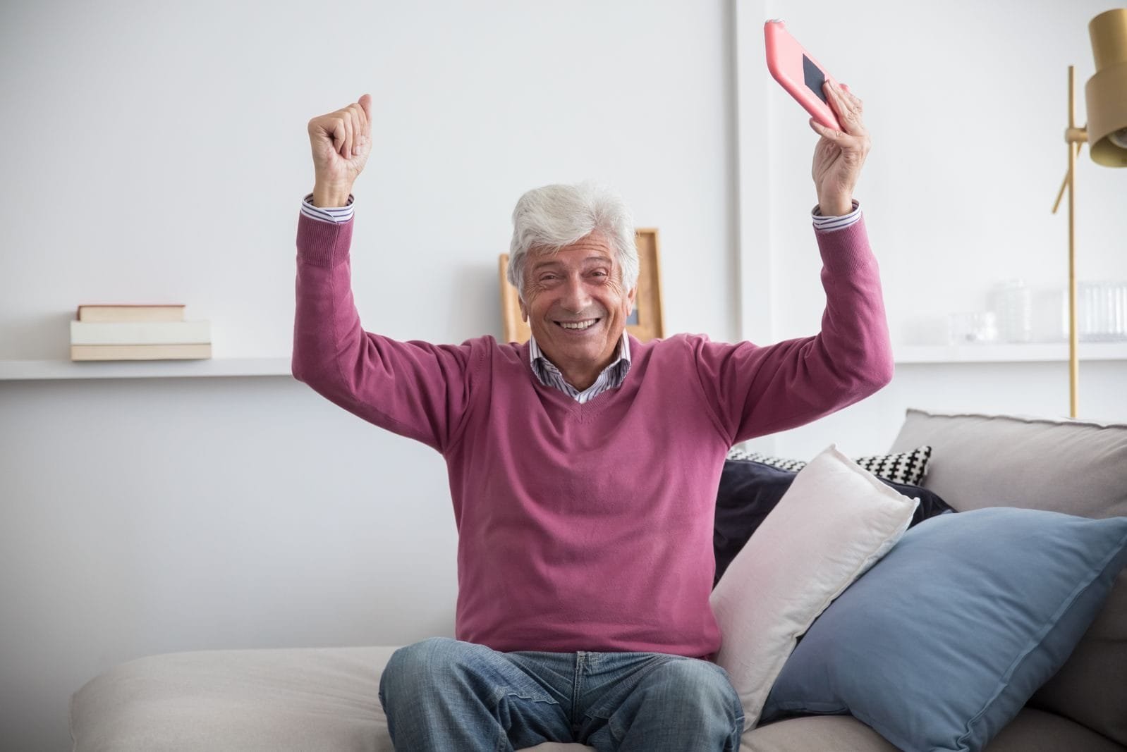 Golpe do App Falso do INSS: Como se Proteger em 2026 1 A happy senior man sitting on a sofa holding a smartphone indoors, celebrating with raised arms.