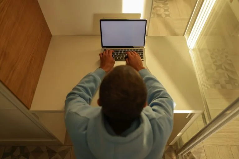 Overhead view of a man typing on a laptop with a blank screen, wearing a hoodie, sitting a