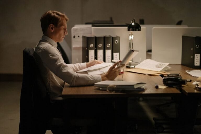An office worker in formal attire intensely focused while reading documents at a desk with folders and a lamp.