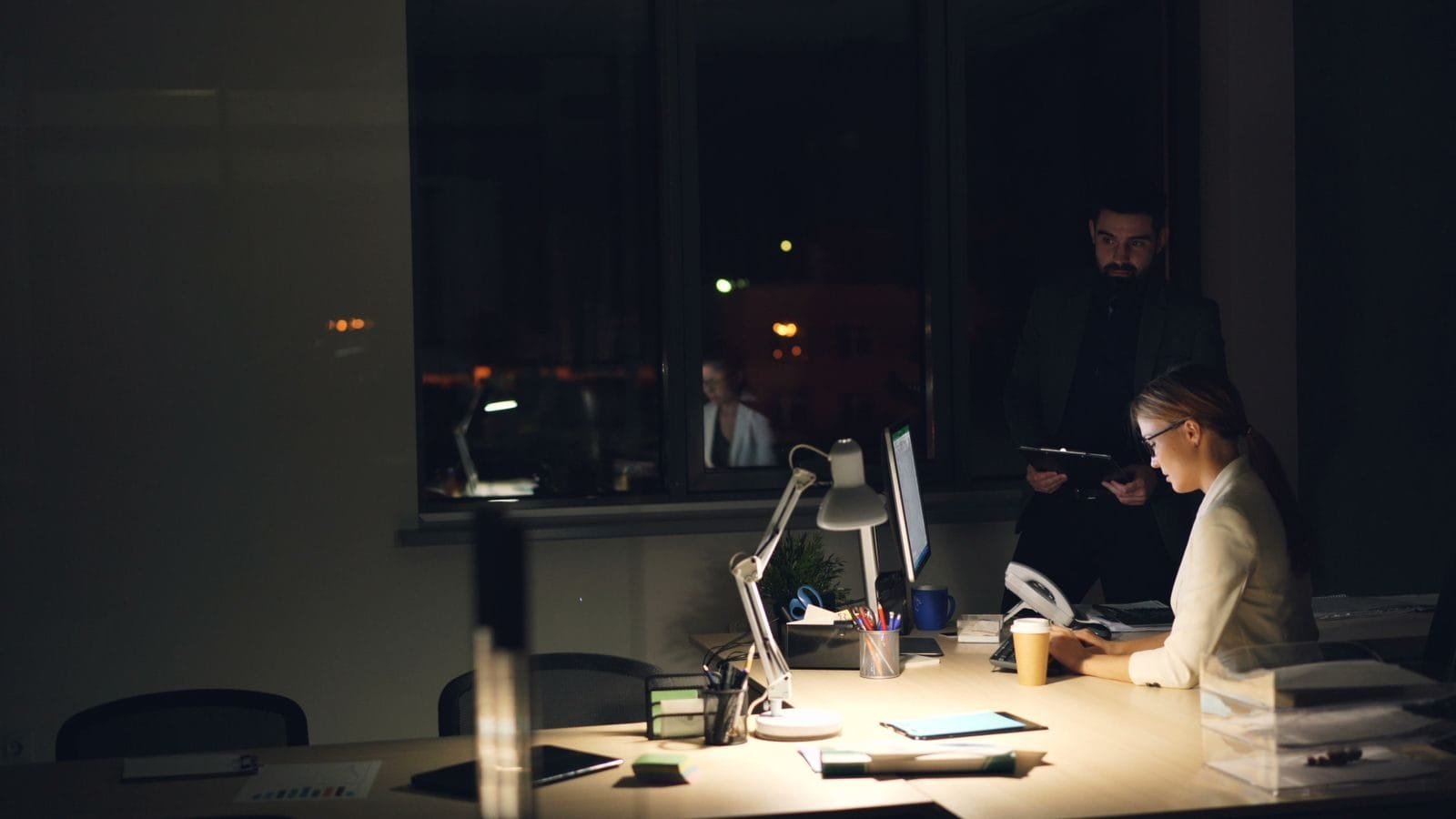 Horas extras do bancário refletem no décimo terceiro e no INSS 1 Two professionals working late in a dimly lit office, focused on tasks.