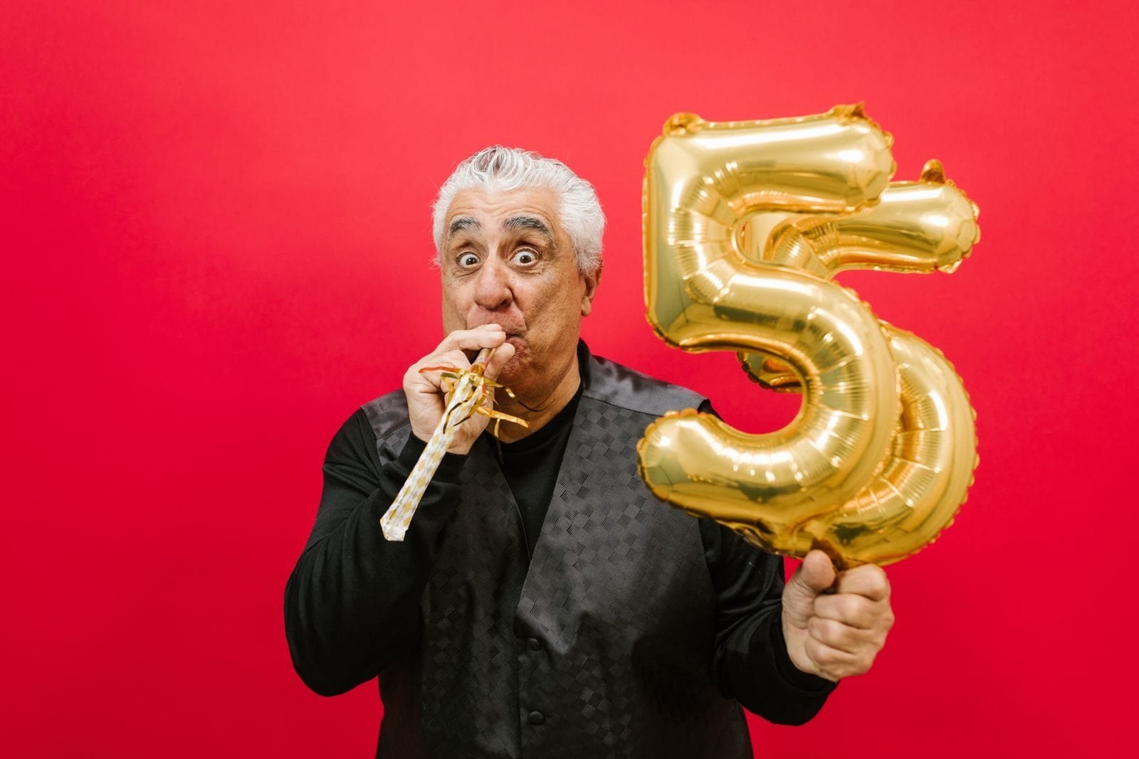 Idade Mínima Progressiva: Tabela Atualizada, Requisitos e Como Calcular 1 Senior man with a party horn and gold balloon, celebrating in a studio with a vibrant red background.