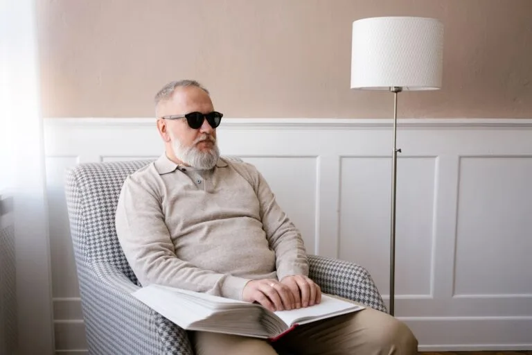 Elderly man with sunglasses reading a braille book indoors, highlighting disability awareness.