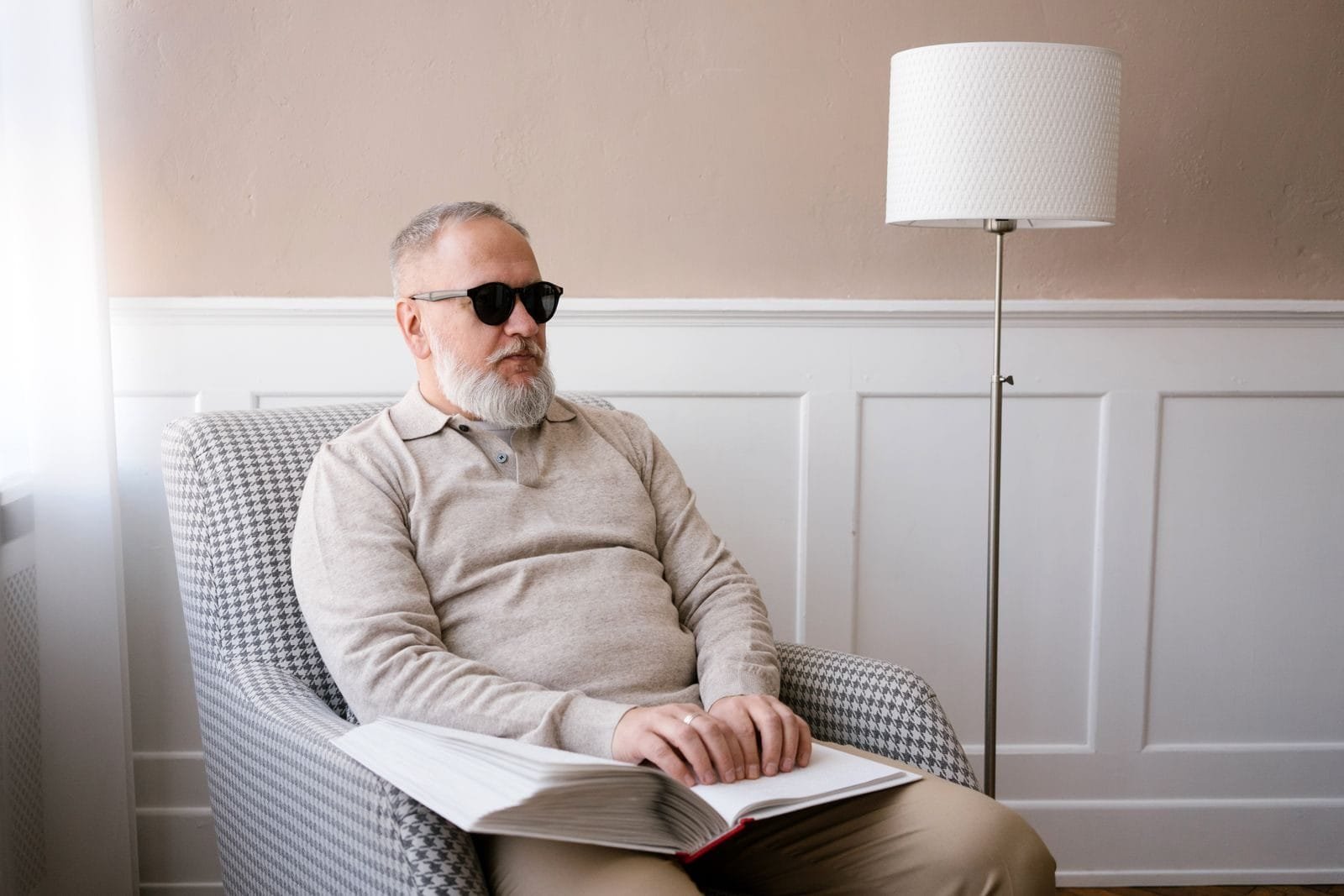 INSS adota fila nacional única para análise de benefícios, com prioridade a incapacidade e BPC 1 Elderly man with sunglasses reading a braille book indoors, highlighting disability awareness.