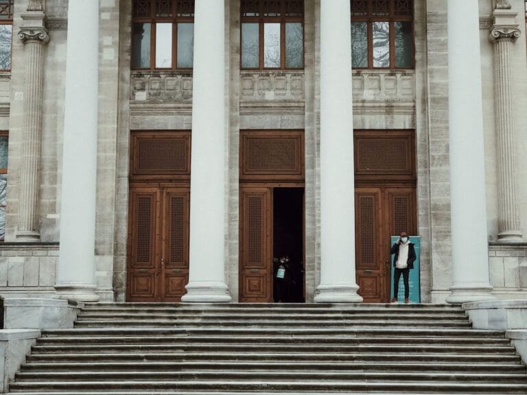 Exterior view of a neoclassical building with large columns and a staircase, featuring a person entering.