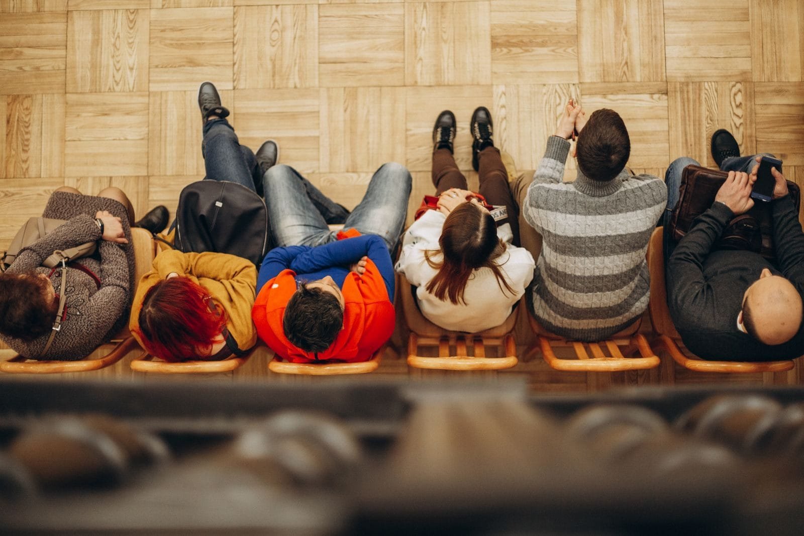 Top view of adults sitting in a row on wooden chairs on a parquet floor indoors.