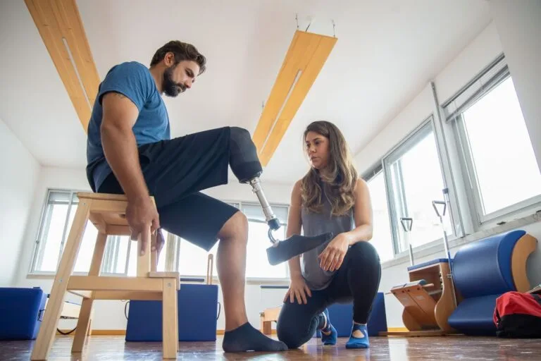 INSS Entrega Próteses a Segurados em Reabilitação em 2026 4 A man with a prosthetic leg assisted by a therapist during rehabilitation indoors.