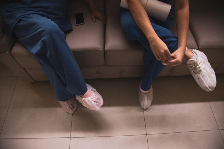INSS faz mutirão com quase 19 mil perícias médicas antecipadas em 92 agências 12 Two healthcare workers sitting on a sofa wearing scrubs and protective shoe covers.