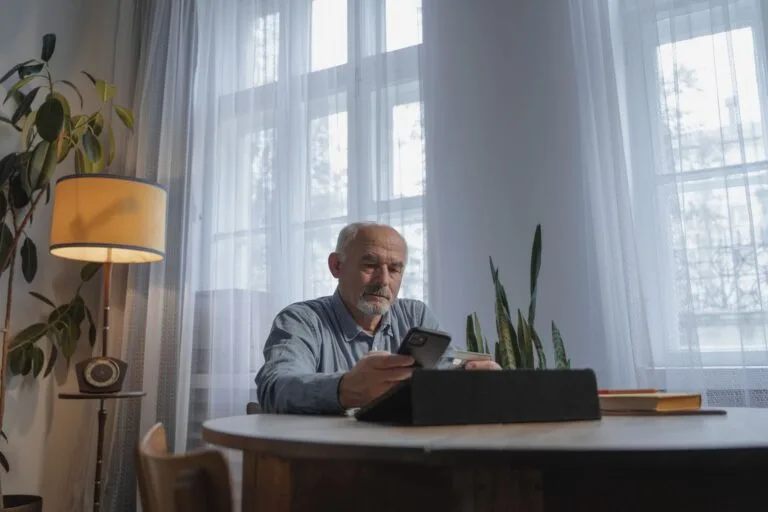 Elderly man with facial hair using phone at home desk, engaging in online activities in a comfortable setting.