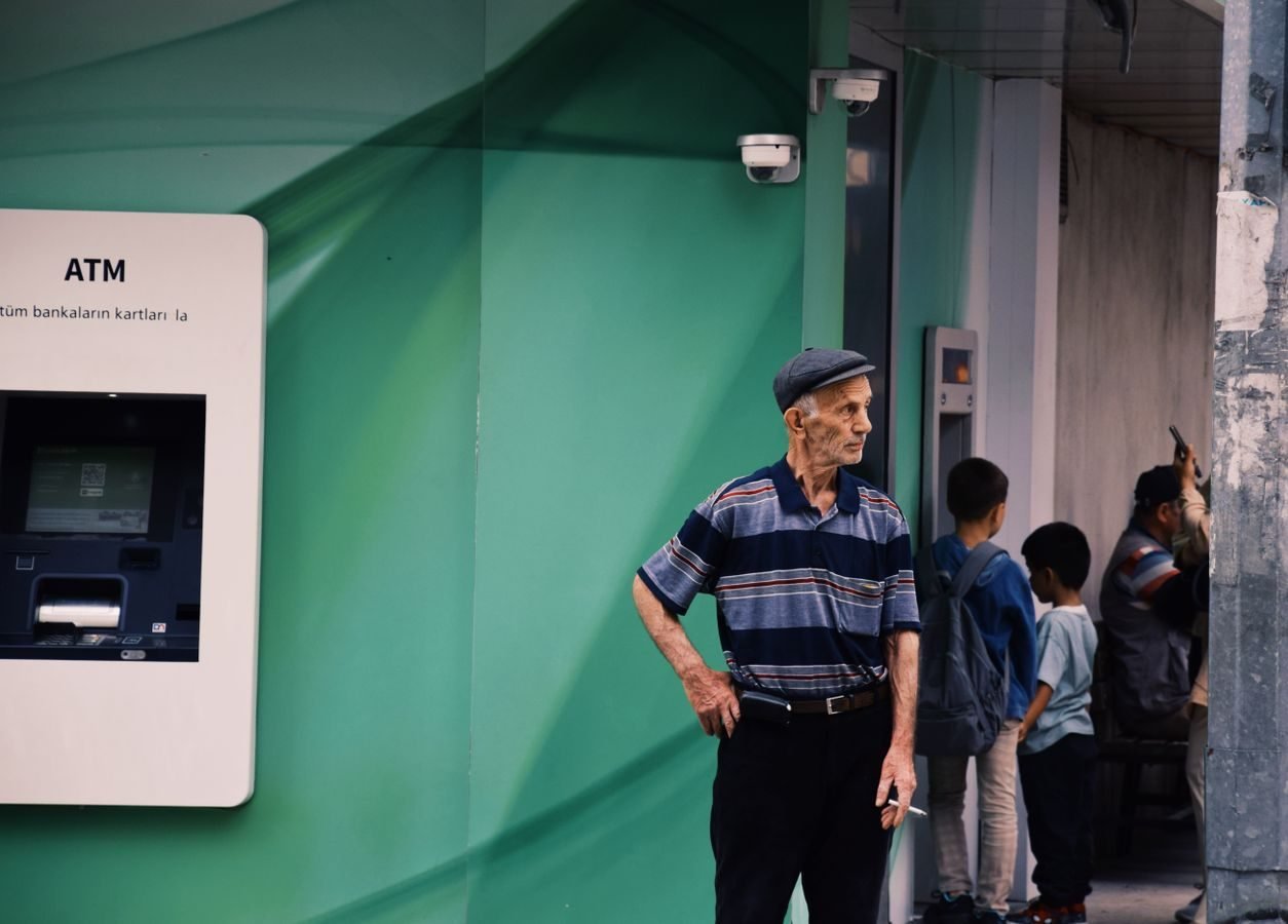 INSS inicia em 24 de abril de 2026 o pagamento da primeira parcela do 13º salário antecipado a aposentados e pensionistas 1 Elderly man in a striped shirt stands by an ATM with children nearby in an urban setting.
