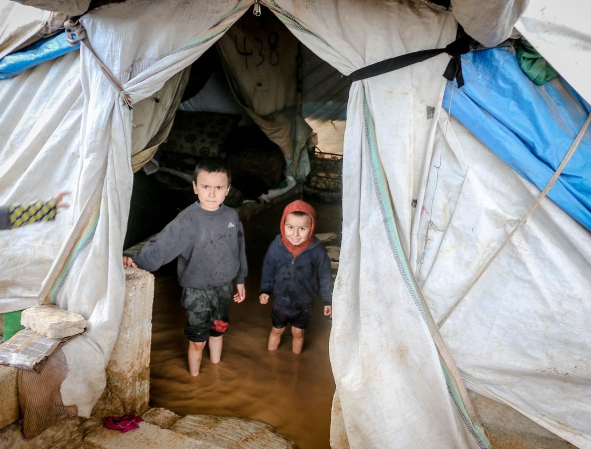 Two young children stand in a flooded tent in Idlib, Syria, highlighting the hardships in refugee camps.