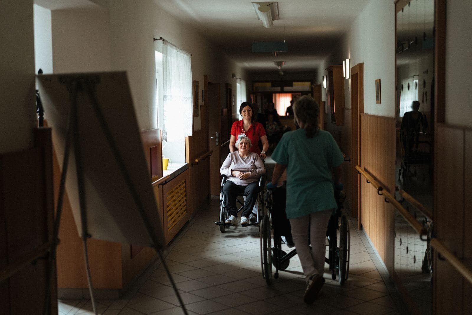 INSS Supera 620 Atendimentos no Governo na Rua em Macapá 1 Caregiver assisting elderly woman in a wheelchair through a retirement home corridor.
