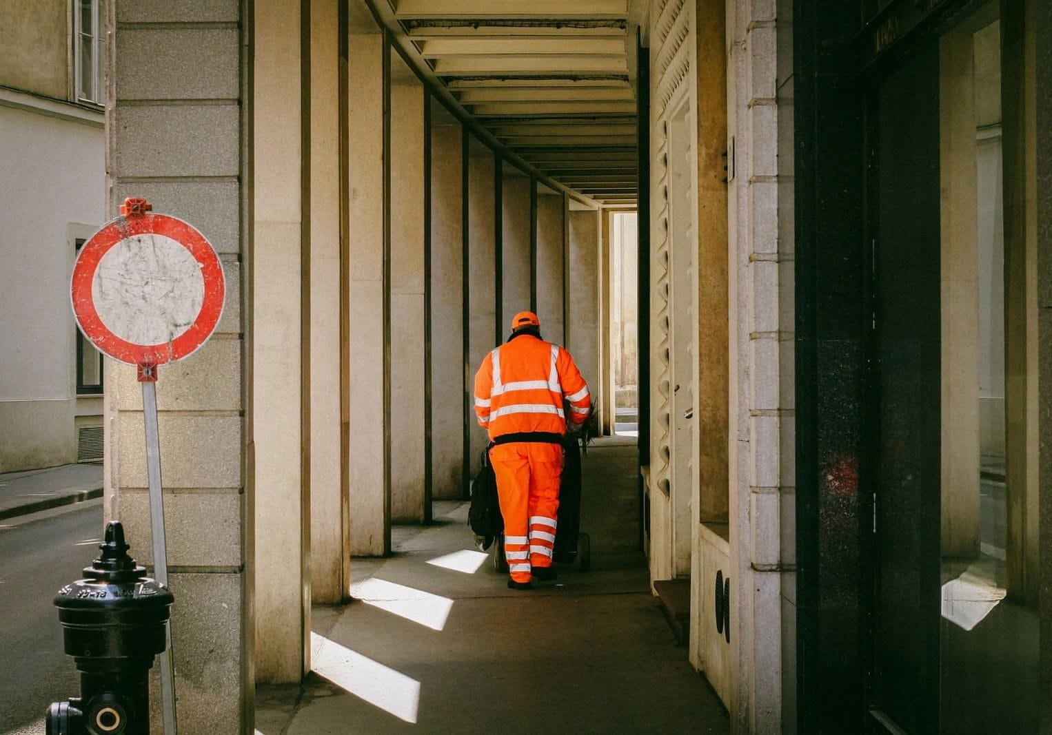 ISS: como saber em qual município o imposto sobre serviços é devido 1 Street worker in orange uniform pushing a cart down a narrow urban alley during the day.