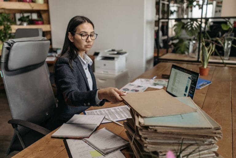 Como podemos ajudar? 15 Businesswoman working at a wooden desk with documents and laptop in a modern office.