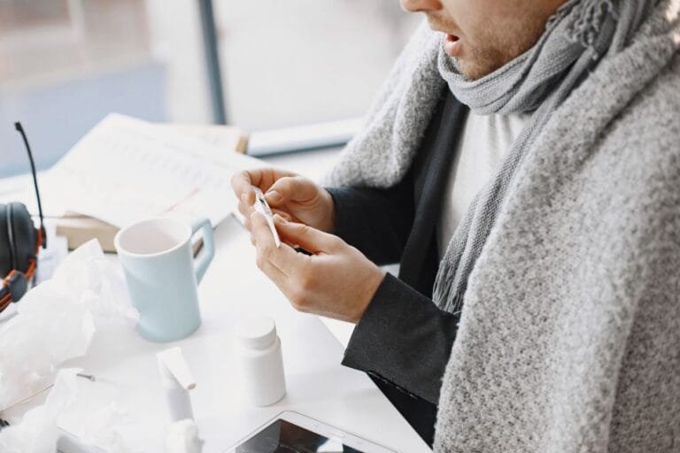 Como podemos ajudar? 12 A man wrapped in a blanket, checking a thermometer while convalescing indoors.