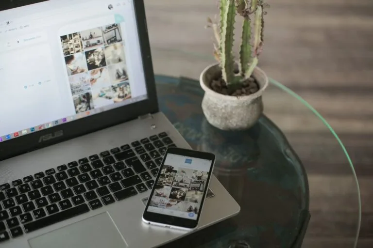 A laptop and smartphone displaying social media apps next to a cactus on a glass table.