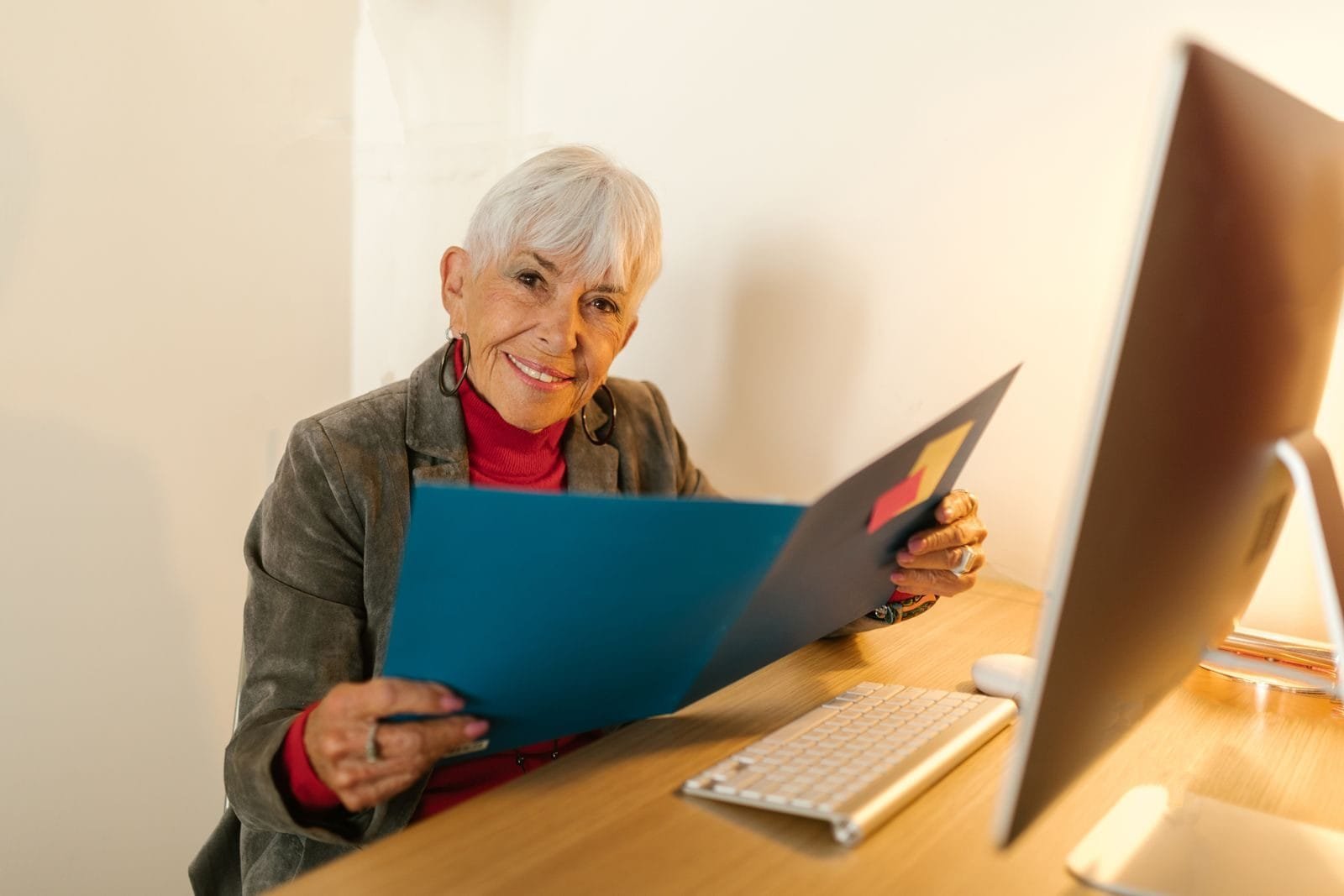 Margem consignável do INSS terá redução gradual de 2 pontos percentuais ao ano, chegando a 30% em 2031, conforme regra divulgada em maio de 2026 1 Elderly woman in business attire sitting at desk, holding a folder and smiling warmly in an office setting.