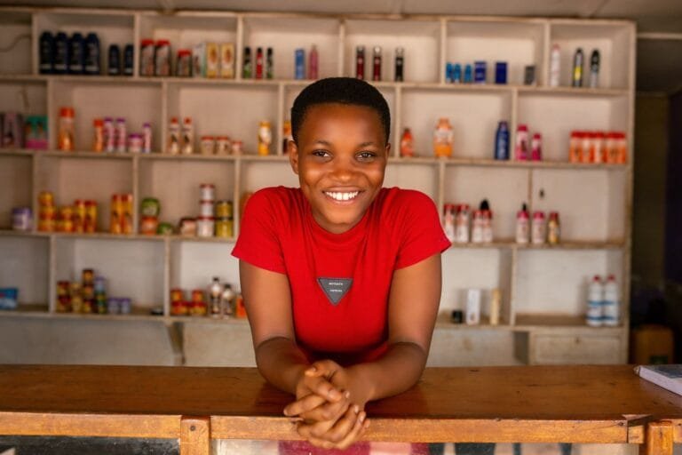 A cheerful woman stands behind a wooden counter, smiling in front of stocked shelves in a store.