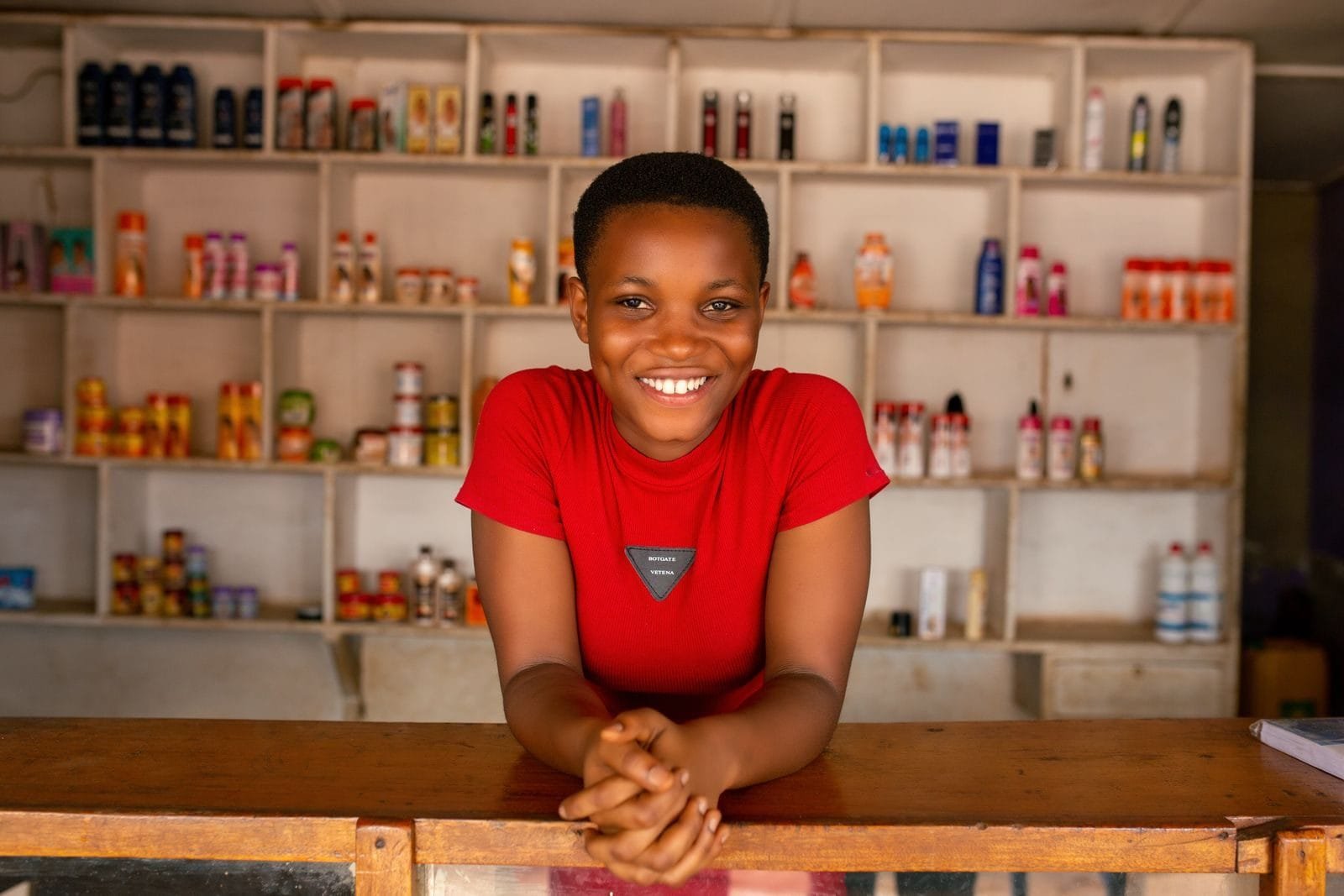 A cheerful woman stands behind a wooden counter, smiling in front of stocked shelves in a store.