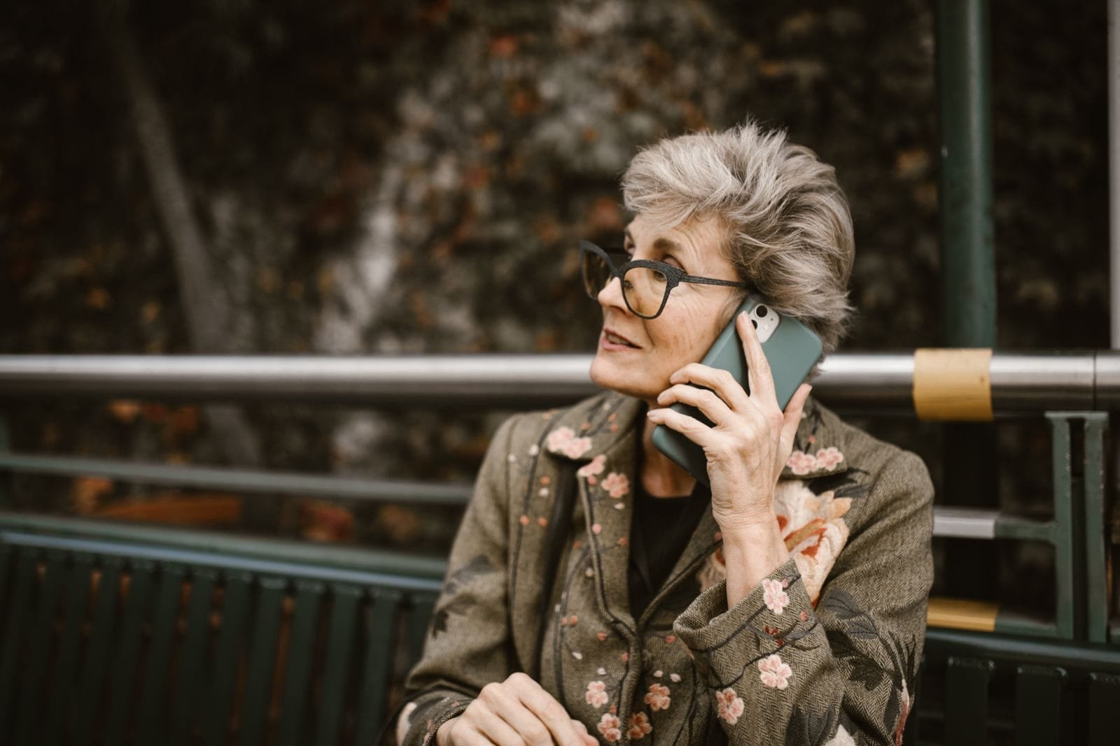 Elderly woman with glasses talking on a smartphone while seated outdoors.