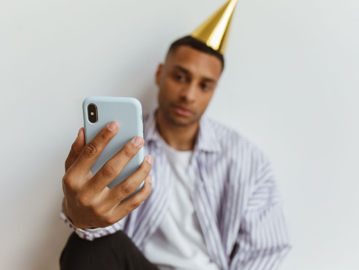 Meu nome esta sendo usado em perfil falso nas redes sociais 1 A man wearing a party hat takes a selfie with his smartphone indoors against a plain white background.