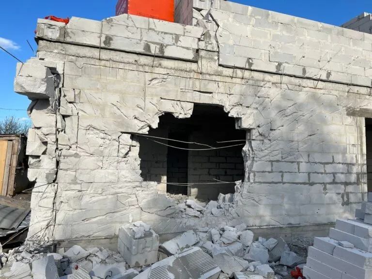 Concrete building wall with a large hole, showing damage and rubble, under a clear blue sky.