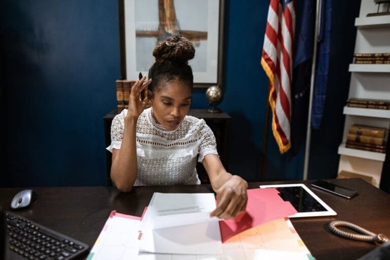 Como podemos ajudar? 4 Businesswoman examines reports at desk with American flag in background.