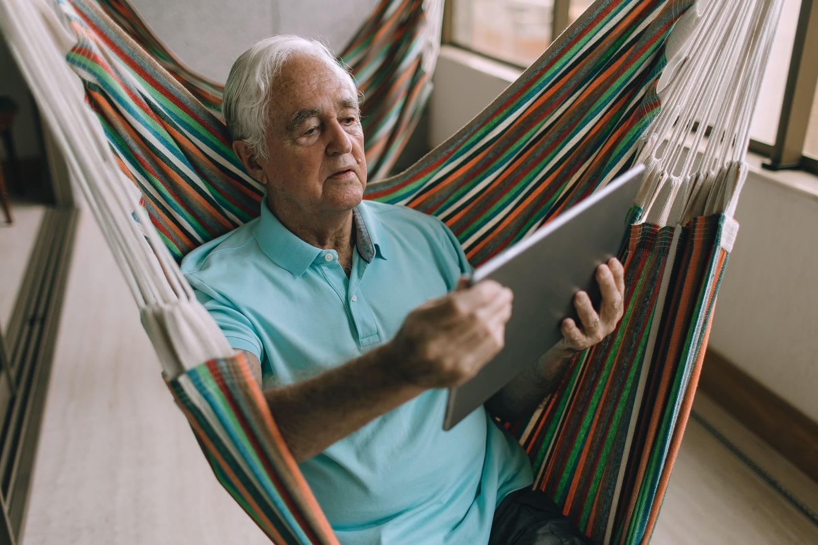 Pedágio de 100%: Cálculo e Vantagens Sobre Outras Regras 1 Elderly man using a tablet while relaxing on a hammock on a balcony.