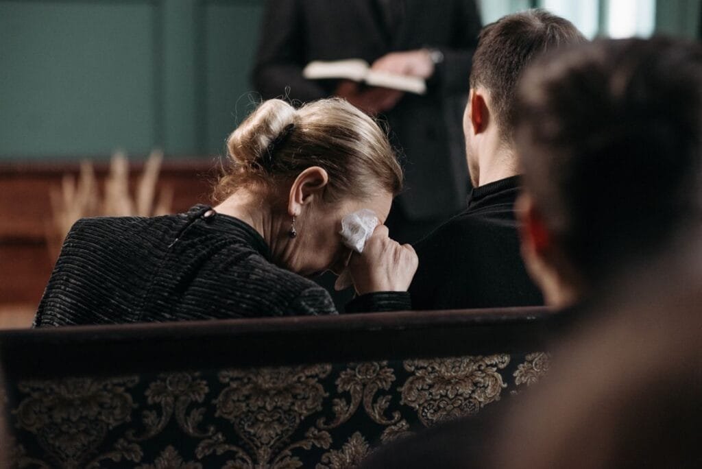 Meu marido faleceu. Tenho direito a pensão por morte? 1 A grieving woman wipes tears during a heartfelt funeral ceremony in a somber indoor setting.