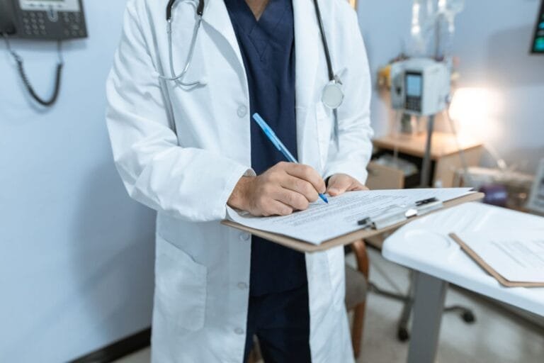 Portaria Conjunta MPS/INSS nº 13/2026 cria o Novo Atestmed e amplia o prazo da análise documental de 60 para até 90 dias 7 A doctor in a lab coat writing notes in a hospital setting, using a clipboard.