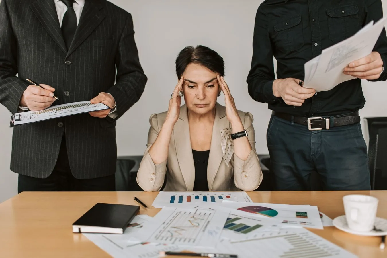 Pressão para vender seguros e consórcios e o adoecimento bancário 1 A stressed businesswoman holding her head with colleagues holding documents in a corporate office setting.